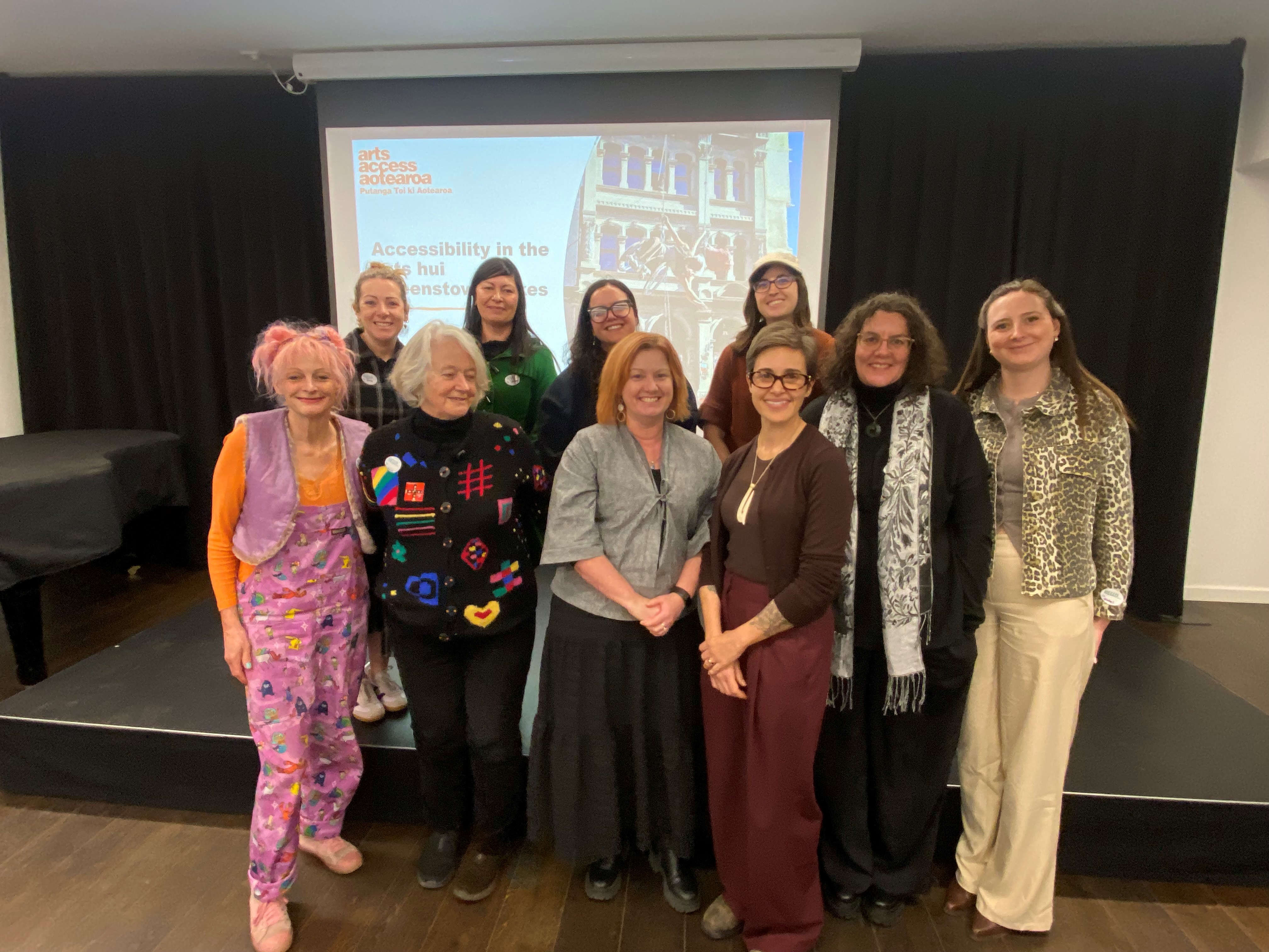A group of ten women stand together in front of a projector screen at the Queenstown Arts Accessibility hui 2026